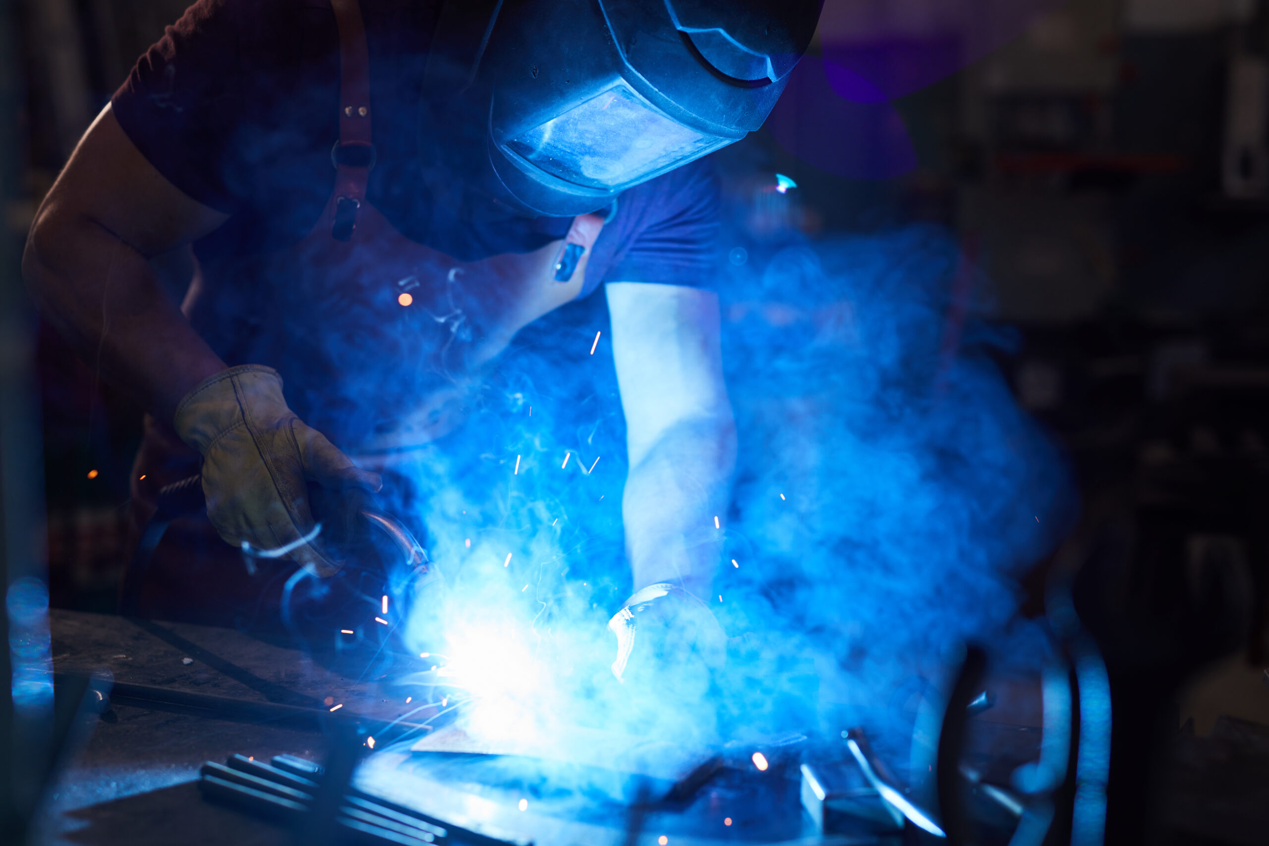 Busy mechanic in work helmet standing at desk and using welding torch while working with metal in smithy